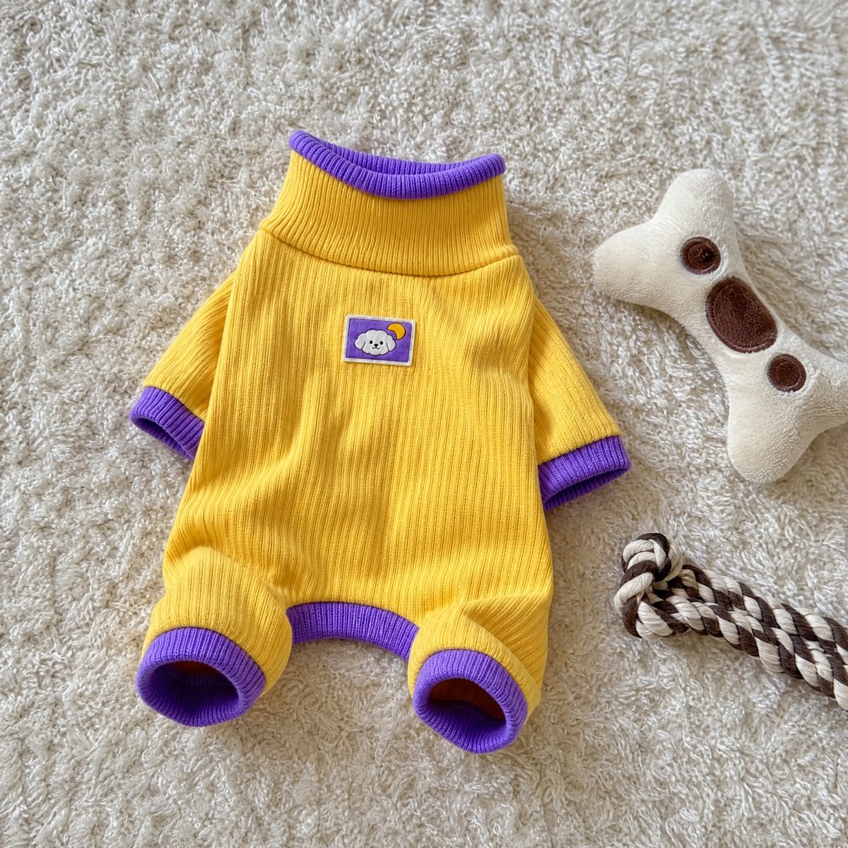Ein gelber, gerippter Hundeoverall mit lila Rollkragen und Bündchen liegt auf einem weißen Teppich neben Hundespielzeug.
