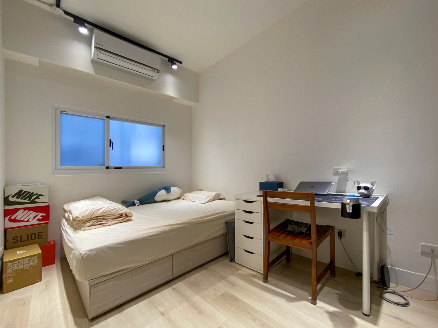 Small bedroom before: bed with cream bedding, white desk with drawers and a brown wooden chair, shoe boxes stacked on the left, sliding window, air conditioner and ceiling track light