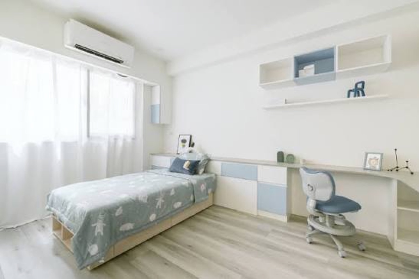 Kids' bedroom before: single bed with blue star-pattern bedding, built-in white desk with blue drawers and wall shelves, a blue desk chair, a wide sliding window on the left wall and an air conditioner