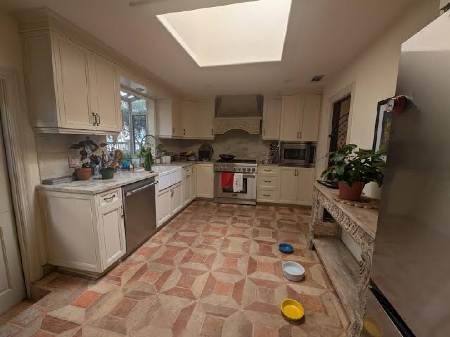 Kitchen before: cream cabinets, terracotta patterned floor tile, a stainless range and hood, a skylight, plants on the counter and pet bowls on the floor